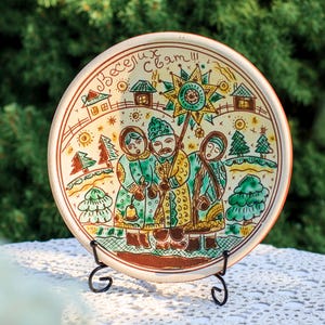 Puede incluir: Plato redondo de cerámica pintado a mano con un diseño decorativo de arte popular. El plato presenta una imagen central de tres figuras con vestimenta tradicional, rodeadas de casas, árboles y una estrella. El borde tiene un borde marrón y el plato se exhibe sobre un soporte de metal negro.