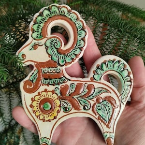 Op de afbeelding: Een crème-kleurige keramische ramfiguur, versierd met bruine, groene en gele bloemen- en krulwerkpatronen. De ram heeft grote, spiraalvormige hoorns. Het beeldje wordt tegen een achtergrond van groen gebladerte gehouden.