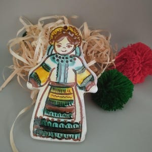 Op de afbeelding: Handbeschilderde keramische figuur van een vrouw in traditionele klederdracht. Het beeldje heeft een witte basis met een blauwe, gele en bruine jurk. De vrouw heeft bruin haar en is omgeven door decoratief stro en twee pompons.