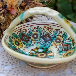 Op de afbeelding: Een handbeschilderde keramische mand met een handvat. De mand heeft een crèmekleurige basis met een bloemenontwerp in teal, geel en bruin. Het handvat heeft teal en gele accenten. De mand staat op een wit kanten tafelkleed.