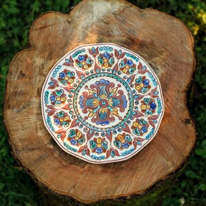 Peut inclure: Une assiette décorative en céramique avec un motif floral détaillé dans les tons de bleu, jaune et rouge. L'assiette est posée sur une surface en bois rustique, mettant en valeur ses motifs complexes et ses couleurs vives. Le diamètre de l'assiette est d'environ 20 cm.