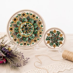 Puede incluir: Dos platos redondos de cerámica con un patrón detallado estilo mandala en tonos verde, amarillo y marrón. El plato más grande a la izquierda y el más pequeño a la derecha muestran motivos florales y geométricos sobre una base crema.