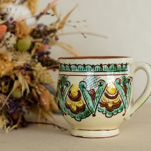 May include: A ceramic mug with a cream-colored base and a decorative handle. The mug features a hand-painted design with green, yellow, and brown floral motifs. A bouquet of dried flowers is in the background.
