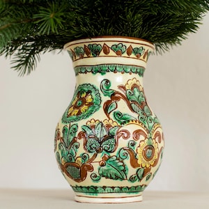 Puede incluir: Un jarrón de cerámica color crema con un diseño floral detallado en tonos verdes, marrones y amarillos. El jarrón está lleno de ramas de pino verdes, creando un contraste natural. El jarrón tiene forma bulbosa y un borde decorativo.