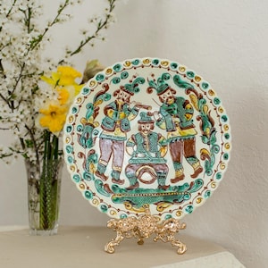 Puede incluir: Un plato decorativo de cerámica con un diseño de arte popular, que presenta tres figuras tocando instrumentos. El plato está sostenido por un soporte de metal dorado. Un jarrón de flores está en el fondo.