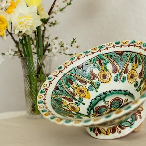 May include: A decorative, hand-painted ceramic bowl with a pedestal base. The bowl features a white background with a floral design in yellow, green, and brown. A vase of daffodils and white flowers is in the background.