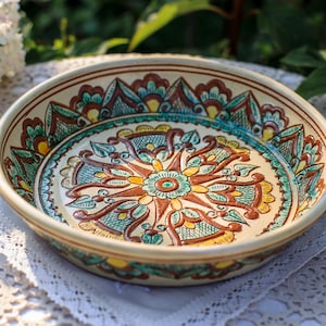 May include: A round, hand-painted ceramic bowl with a decorative floral design. The bowl features a cream-colored base with a detailed pattern in shades of brown, teal, yellow, and green. The bowl is sitting on a white lace doily.