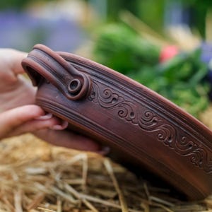 Può includere: Una ciotola in ceramica marrone con manico e decorazioni a volute. La ciotola è tenuta in mano, mostrando il suo design dettagliato. La ciotola viene probabilmente utilizzata per servire il cibo.