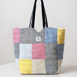 Puede incluir: Un bolso tote colorido con un diseño de patchwork. El bolso presenta una variedad de colores, incluyendo rosa, azul, amarillo y negro, dispuestos en un patrón geométrico. Tiene asas negras y una pequeña etiqueta con un diseño de hoja.