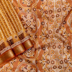 May include: Close-up of a textile with a warm colour scheme. The fabric displays complex patterns in gold, brown, and white. The design incorporates floral and geometric motifs, with a decorative border. The textile is likely a saree or similar garment.