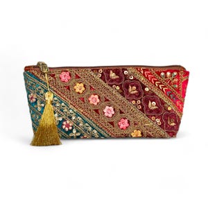 Puede incluir: Un bolso de mano rectangular multicolor con cremallera y borla doradas. El bolso presenta un diseño de patchwork con paneles turquesa, burdeos y rojos, adornado con bordados dorados, lentejuelas y detalles florales.