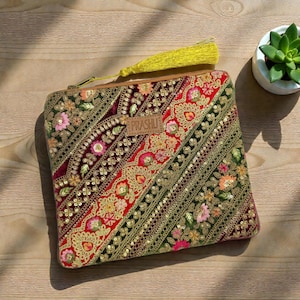 May include: An embroidered clutch with a vibrant design. The clutch features a mix of red, green, and gold colors with floral patterns and sequins. A yellow tassel hangs from the zipper. The word "PRASHU" is on a small brown label.