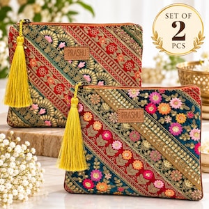 May include: A set of two rectangular pouches featuring intricate embroidery in red, green, and gold with floral patterns and diagonal stripes. Each pouch has a yellow tassel attached to the zipper and a brown leather label with the text PRASHU.