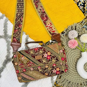 May include: A burgundy and gold embroidered handbag with a matching shoulder strap. The bag features floral and geometric patterns, with a brown leather handle and strap attachments. The strap is approximately 2 inches wide. The bag is set against a colorful background.