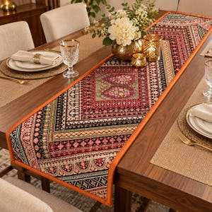 Könnte beinhalten: Ein langer, rechteckiger Tischläufer mit einem komplexen, mehrfarbigen Muster in den Farben Dunkelrot, Schwarz, Gold und Creme. Das Design umfasst komplizierte geometrische Bordüren und florale Motive, eingefasst mit einem einfarbigen orangefarbenen Stoffrand.