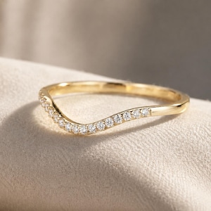 May include: A curved yellow gold band ring featuring a row of small, clear, round-cut stones set along the front arc. The metal has a polished, reflective finish, and the design follows a gentle wave shape that tapers toward the back of the band.