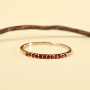 Könnte beinhalten: Ein zarter Goldring mit einer Reihe kleiner, leuchtend roter Edelsteine. Der Ring ist vor einem weichen, neutralen Hintergrund platziert, der das Funkeln der Edelsteine hervorhebt. Das Design des Rings ist schlicht und elegant, geeignet für den täglichen Gebrauch.