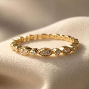 Puede incluir: Un anillo de oro con un patrón alterno de engastes en forma de marquesa y pequeños engastes redondos, cada uno con una piedra clara y brillante. El metal tiene un acabado pulido y el diseño crea un aspecto continuo y delicado.