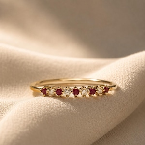 May include: A thin gold band ring featuring an alternating row of four round red rubies and four round clear white diamonds set in a prong setting across the top half of the band, creating a delicate and symmetrical design on the jewellery piece.