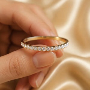 May include: A thin gold band ring featuring a continuous row of small, round, clear white stones set along the front half of the band. The metal has a polished yellow gold finish, and the stones are arranged in a uniform, side-by-side pattern.