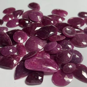 May include: A collection of loose, faceted rubies in various shapes and sizes, including oval, round, and pear cuts. These gemstones exhibit a deep, saturated magenta-red color with visible geometric facets that reflect light across their surfaces.