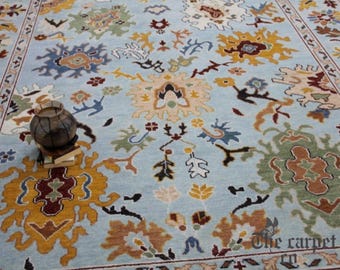 May include: A light blue area rug with an intricate floral pattern in shades of yellow, brown, green, and blue. The rug features a border and a central medallion design. A decorative vase sits on top of the rug.