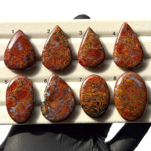 May include: Eight polished cabochon gemstones arranged in two rows on a display tray. Each stone features a complex, swirling pattern of deep red, burnt orange, and muted blue-grey tones. The stones are numbered one through eight.