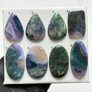 Puede incluir: Ocho piedras preciosas pulidas en cabujón dispuestas en dos filas sobre un tablero de exhibición blanco texturizado. Cada piedra presenta patrones únicos de colores morado, verde, azul y crema, numeradas del uno al ocho para identificación.