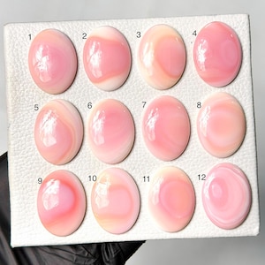 May include: Twelve oval-shaped pink conch shell cabochons arranged in a grid on a white textured display board. Each cabochon features unique swirling patterns of light and dark pink hues with a polished, glossy finish, numbered one through twelve.