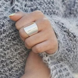 May include: A silver-coloured, wide band ring is worn on a hand, set against a backdrop of a grey, textured sweater. The ring has a smooth, polished surface and a rectangular shape, reflecting light.