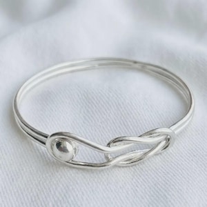 May include: A polished silver bangle bracelet featuring a continuous circular band that loops into a decorative knot design on one side. The metal has a smooth, reflective surface with a bright metallic finish, forming a singular, cohesive piece.