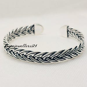 Puede incluir: Un brazalete plateado con un diseño trenzado. El brazalete abierto presenta un patrón tejido, con el texto "kripajewellers21" visible en el lateral. El brazalete se muestra sobre una superficie blanca.