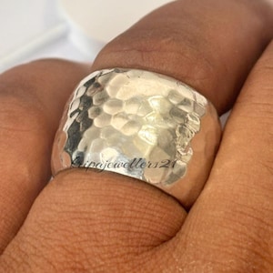 Può includere: Un anello largo in argento con una texture martellata. L'anello è spesso e ha una forma arrotondata. Il testo "ripajewellers21" è inciso sull'anello. L'anello è indossato su un dito.