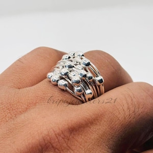 May include: A silver ring featuring multiple bands adorned with small, spherical beads. The ring has a layered design, with the beads clustered at the top. The ring is worn on a finger, showcasing its intricate details and polished finish.