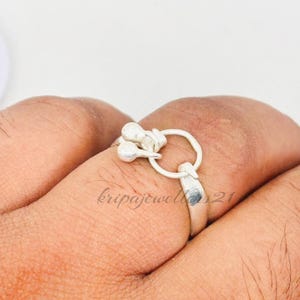 May include: A silver ring with a distinctive design, including a small loop and two spherical ornaments. The ring is worn on a finger, highlighting its intricate details and craftsmanship. The ring is a piece of jewellery.