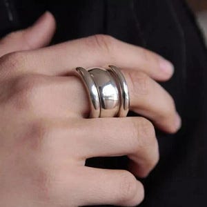 May include: A polished silver ring featuring a central rounded section flanked by two curved bands. The ring's design is contemporary and striking, with a reflective surface. The ring is suitable for everyday wear or special events.