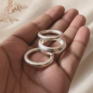 May include: A set of three smooth, polished silver-toned rings with a rounded, thick band profile. The rings are stacked together, showcasing a reflective metallic surface and a simple, circular design without any additional embellishments or stones.