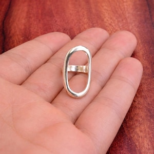 May include: A silver-colored ring with an elongated oval design. The ring features a band that connects the top and bottom portions of the oval. The ring is shown on a hand against a brown background.