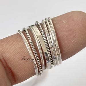 Puede incluir: Una colección de anillos de plata en varios diseños, incluyendo bandas martilladas, trenzadas y lisas. Los anillos están apilados, mostrando diferentes texturas y estilos. Los anillos están hechos de un material metálico brillante.