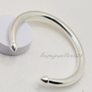 May include: A silver cuff bracelet with a rounded, smooth design. The bracelet has pointed ends and is displayed on a white surface. The jewellery piece is a simple, elegant accessory.
