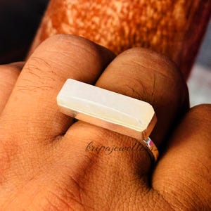 May include: A rectangular, pale gemstone ring with a silver band. The gemstone has a smooth, polished surface. The ring is shown on a person's finger. The text "kripajewellery" is visible on the ring.