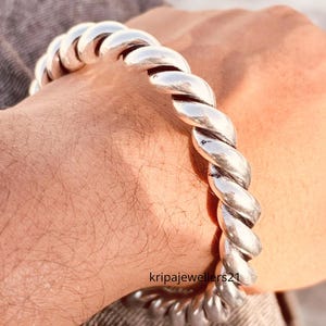 May include: A silver-colored, twisted rope design bracelet. The bracelet is thick and appears to be made of metal. The bracelet is worn on a wrist. The text "kripajewellers21" is visible.