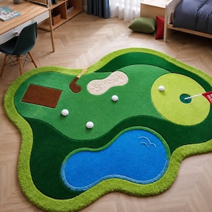May include: An irregularly shaped area rug designed as a miniature golf course. The rug features shades of green grass, a blue water hazard, a tan sand bunker, white golf balls, a brown club, and a red flag with the text "MINI GOLF 1".