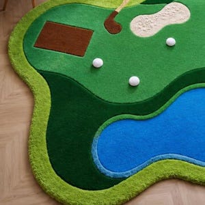 May include: A tufted area rug shaped like a golf course, featuring a vibrant green fairway, a blue water hazard, a brown rectangular tee box, a beige sand bunker, a brown golf club, and three white golf balls scattered across the surface.