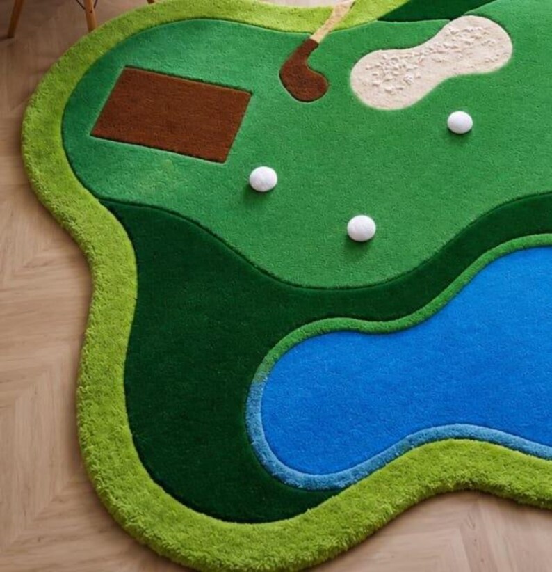 May include: A tufted area rug shaped like a golf course, featuring a vibrant green fairway, a blue water hazard, a brown rectangular tee box, a beige sand bunker, a brown golf club, and three white golf balls scattered across the surface.