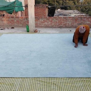 Puede incluir: Una alfombra grande, cuadrada y de color azul claro con una superficie lisa y sólida. El texto SHIMPI RUGS es visible en la esquina superior izquierda. La alfombra está extendida sobre una esterilla de rejilla amarilla y negra en un tejado.