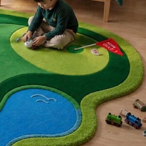 May include: A large, irregularly shaped area rug designed to look like a golf course, featuring shades of green, a blue water hazard, a beige sand trap, and a red flag with the text GOLF 1. The rug has a soft, textured pile surface.