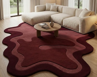 May include: A burgundy and rose-colored area rug with an organic, flowing design. The rug is placed on a light wood floor, beneath a beige sectional sofa and a round wooden coffee table. The rug's unique shape adds a modern touch to the living room.