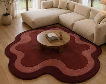 May include: A large, irregular, organic-shaped area rug featuring concentric layers of deep burgundy and muted rose-pink tones. The rug has a soft, textured surface and a wavy, scalloped edge, creating a layered, fluid appearance on the floor.