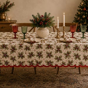 Peut inclure: Une nappe rectangulaire ornée d'un motif répétitif de feuilles de houx vertes et de baies rouges sur fond crème, terminée par une bordure festonnée rouge. La table est dressée avec deux assiettes blanches, des verres rouges et verts, et des bougies.